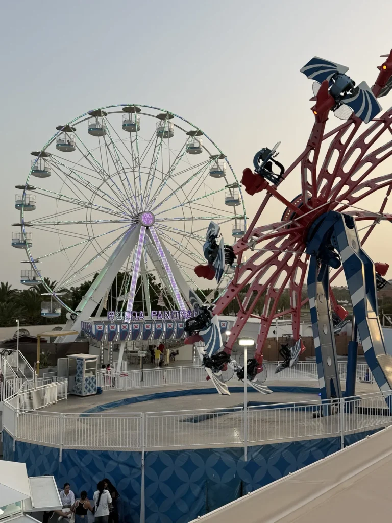 La ruota panoramica illuminata di Holiday World Maspalomas al tramonto, accanto a una giostra rossa e blu